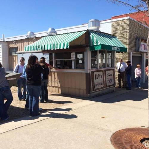 A small hamburger stand with a green and white striped awning, people waiting in line outside on a sunny day.