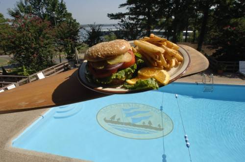 A plate with a burger, lettuce, tomato, and fries sits near a pool with a logo, surrounded by trees and a lake view.
