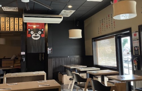 Interior of a restaurant featuring wooden tables, black walls, and a Kumamon bear flag. Bright lighting and large windows.