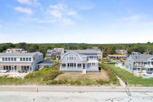 Aerial view of beach houses along a sandy shore, surrounded by greenery and blue skies.