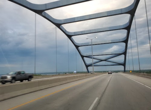 A modern bridge with a unique design spans a highway under a cloudy sky, with vehicles traveling in both directions.