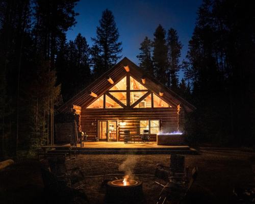 A cozy log cabin at night, illuminated by warm light, surrounded by trees and a glowing campfire in front.