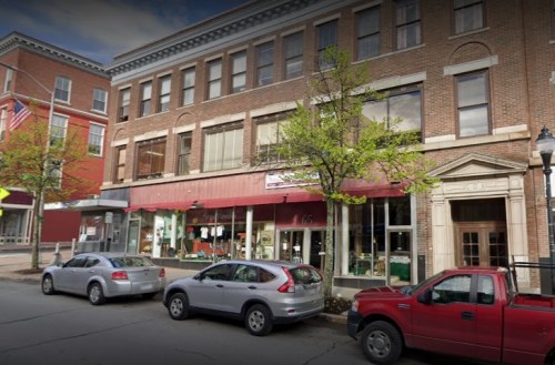 A brick building with large windows and a red awning, flanked by parked cars on a city street.