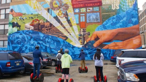 A group of people on segways facing a colorful mural depicting hands and landscapes on a building wall.