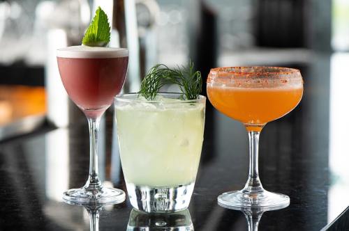 Three colorful cocktails on a dark countertop: a pink drink with mint, a green drink with herbs, and an orange drink with a rim.