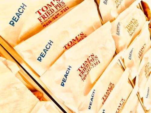 A close-up of paper bags labeled "Tom's Fried Pies" with flavors like "Peach" and contact information visible.