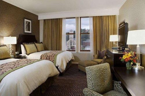A hotel room with two beds, a chair, a desk, and large windows showing a city view. Decorated in warm tones.