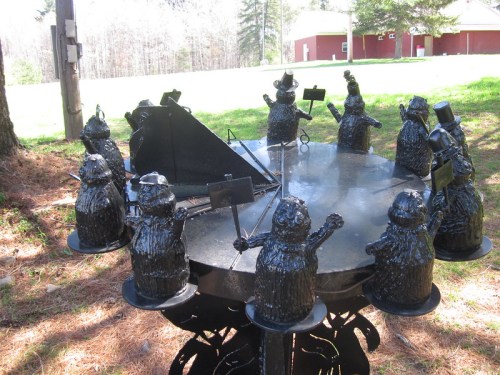 A round table surrounded by whimsical black bear sculptures, each holding signs, in a wooded outdoor setting.