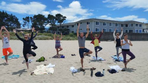A group of people practicing yoga on a beach, balancing in tree pose with a cloudy sky and buildings in the background.