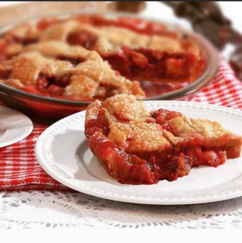A slice of cherry pie on a white plate, with a whole pie in the background on a red checkered tablecloth.