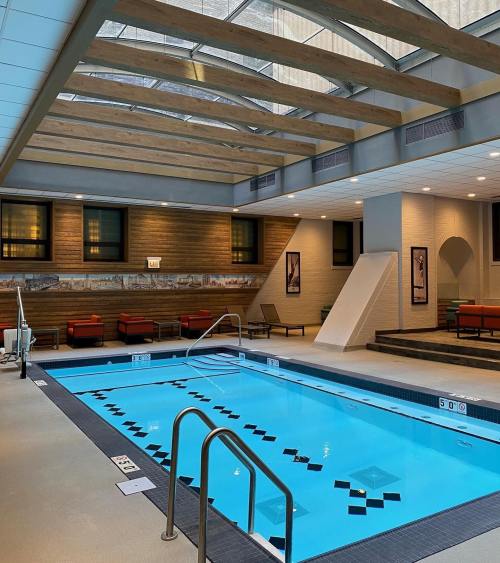 Indoor swimming pool with a glass ceiling, wooden beams, and lounge seating around the edges.