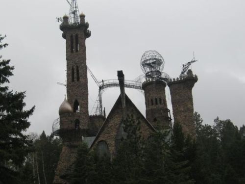 A unique, tall stone castle with towers and antennas, surrounded by dense trees under a cloudy sky.