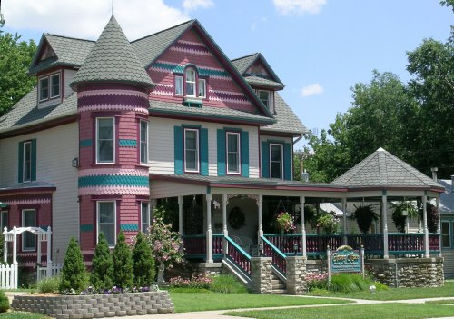 A colorful Victorian-style house with intricate trim and a wraparound porch, surrounded by greenery.