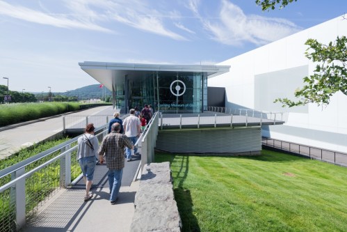 Visitors walking towards the Corning Museum of Glass on a sunny day, with green grass and blue skies in the background.