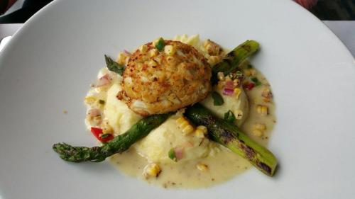 A plate of creamy mashed potatoes topped with a crab cake and grilled asparagus, garnished with corn and vegetables.