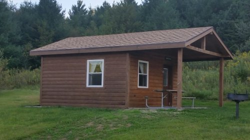 A small wooden cabin with a porch, surrounded by grass and trees in a natural setting.