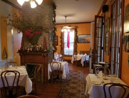 Cozy restaurant interior with wooden floors, tables set with white linens, and festive holiday decorations.