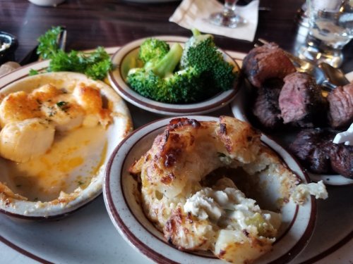 A plate featuring baked seafood, broccoli, and a baked potato with a portion missing, alongside pieces of steak.