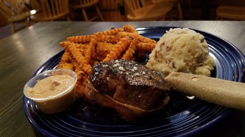 A plate with a grilled steak, sweet potato fries, mashed potatoes, and a small cup of sauce.