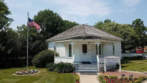 A small white house with a gray roof, surrounded by greenery and flowers, featuring an American flag in front.