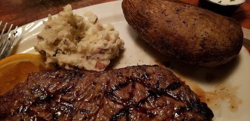 A plate featuring a grilled steak, baked potato, and creamy mashed potatoes, garnished with an orange slice.