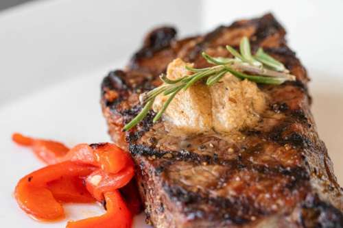 Grilled steak topped with herb butter, garnished with rosemary, and served with roasted red peppers on a white plate.