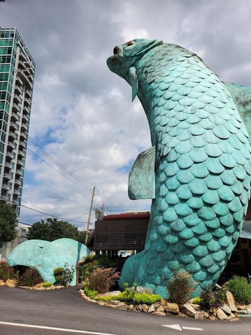 A large, turquoise fish sculpture stands prominently beside a road, with a modern building in the background and cloudy skies.