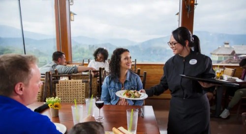 A waitress serves a meal to a smiling woman at a restaurant with a scenic mountain view. Other diners are visible in the background.