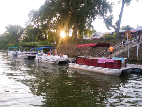 Boats docked along a river at sunset, with people enjoying the evening and trees lining the shore.