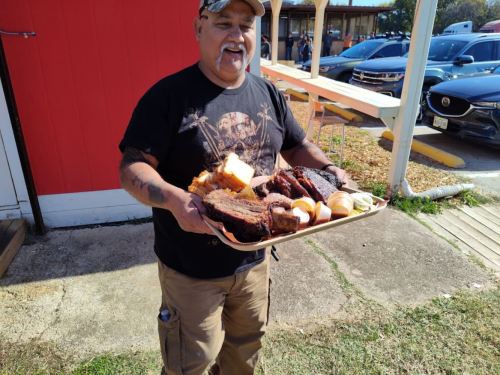 A man holds a large tray of barbecue meats and bread outside a restaurant.