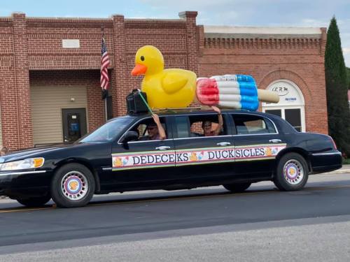 A black limousine with a large rubber duck and popsicle decoration, driving in a parade.