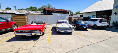 A row of classic cars parked outside a building, including a red convertible, a black and white coupe, and a pink car.