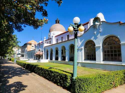 Historic building with a dome, surrounded by greenery and a clear blue sky, featuring arched windows and a walkway.