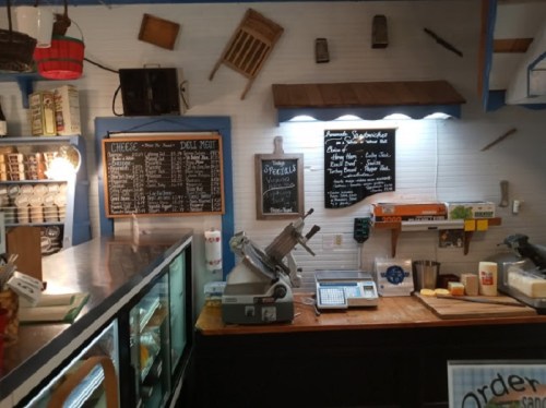 A cozy deli counter with a menu board, meat slicer, and various food items displayed in a rustic setting.