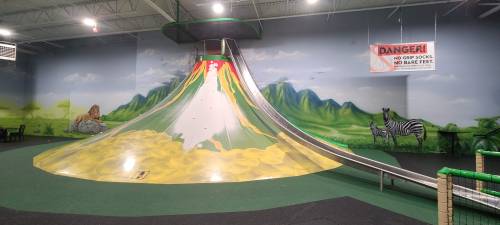 Indoor play area featuring a colorful slide and a large, volcano-shaped climbing structure with jungle-themed murals.