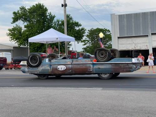 A vintage car with its wheels on top drives past a tent and spectators, showcasing a unique, upside-down design.