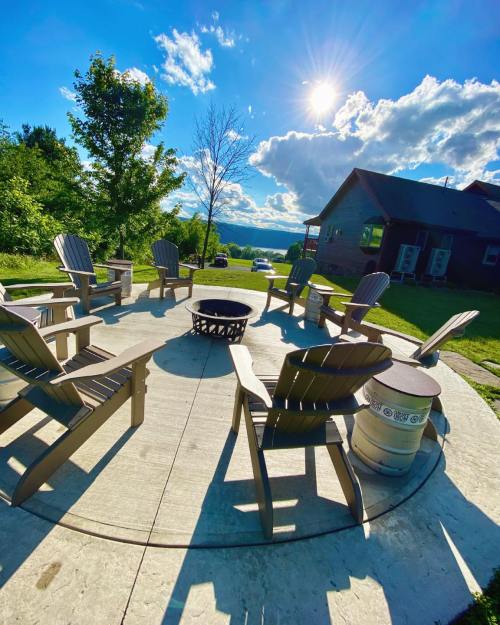A circular patio with wooden chairs surrounding a fire pit, set against a sunny sky and green landscape.