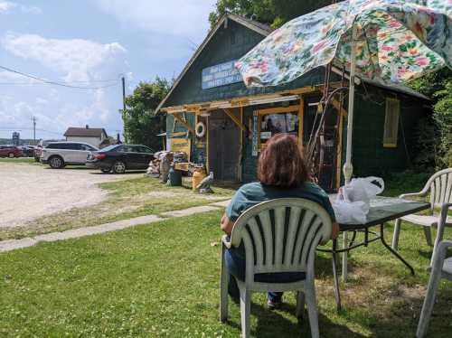 A woman sits under a floral umbrella outside a green building, with cars parked nearby and a grassy area in front.