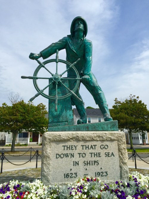 A bronze statue of a sailor at a ship's wheel, commemorating those who went to sea, with flowers in the foreground.