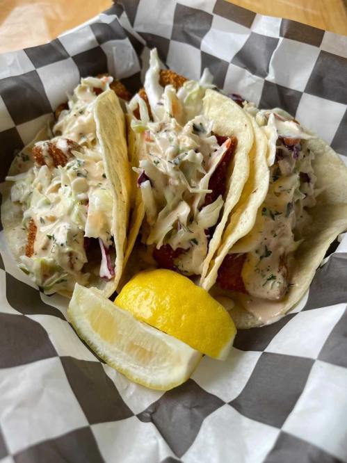 Three fish tacos topped with creamy coleslaw, served with a lemon wedge on a checkered paper plate.