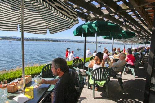 Outdoor dining area by the water, with patrons enjoying meals under green umbrellas on a sunny day.