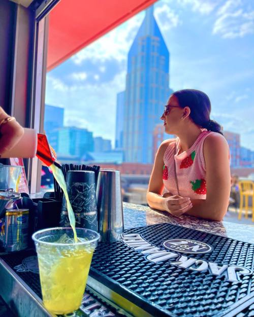 A woman in a pink sweater with strawberries sits at a bar, gazing at a city skyline with a drink being poured.
