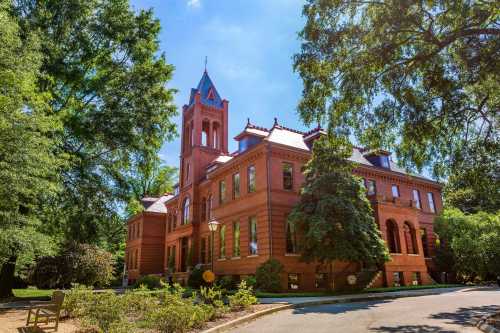 Historic red brick building with a tower, surrounded by lush greenery and a clear blue sky.