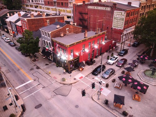 Aerial view of a vibrant street corner with a red building, parked cars, and outdoor seating under umbrellas.