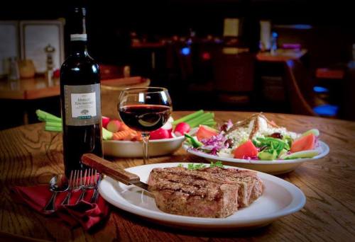 A plate of grilled steak with a glass of red wine and a side salad on a wooden table in a restaurant setting.