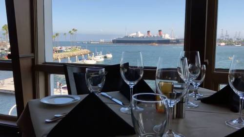 A dining table set with glasses and napkins, overlooking a harbor with boats and the Queen Mary ship in the distance.
