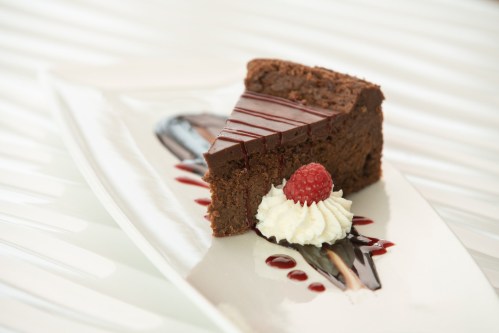 A slice of chocolate cake topped with chocolate drizzle, whipped cream, and a raspberry on a decorative plate.