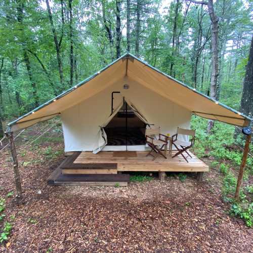 A canvas tent on a wooden platform in a forest, with two chairs on the porch and trees surrounding it.