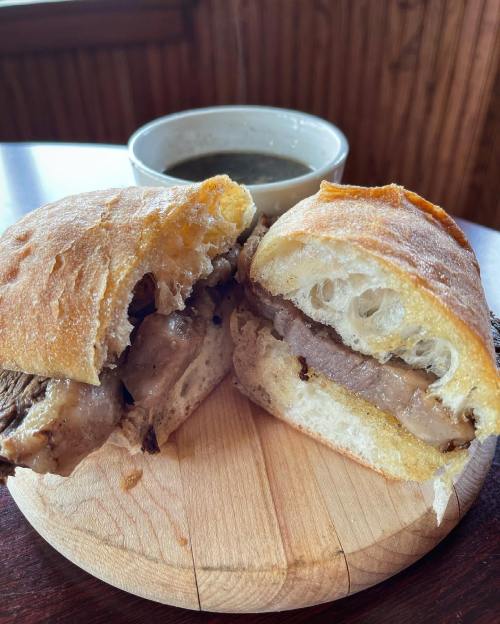 A close-up of a sliced sandwich with meat on a wooden board, accompanied by a bowl of soup in the background.