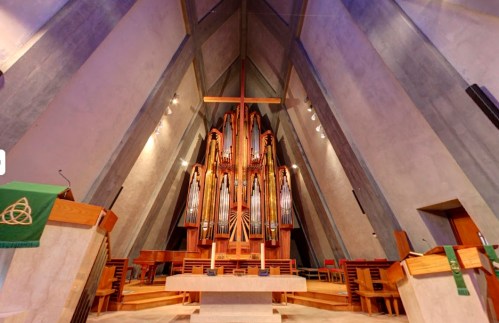 Interior of a modern church featuring a large pipe organ, wooden furnishings, and a tall, angular ceiling.